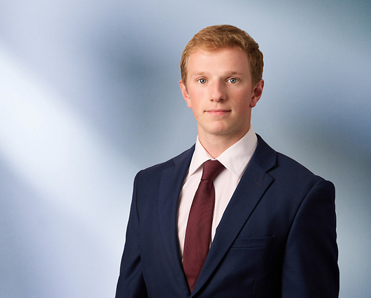 A man, Charles Sarchet, in a dark suit, white shirt, and maroon tie is looking at the camera, standing against a blurred blue and white background.