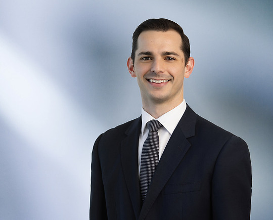 man, Lino Taormina, wearing a dark suit, white shirt, and patterned tie, standing against a softly blurred blue and white background, smiling confidently at the camera.