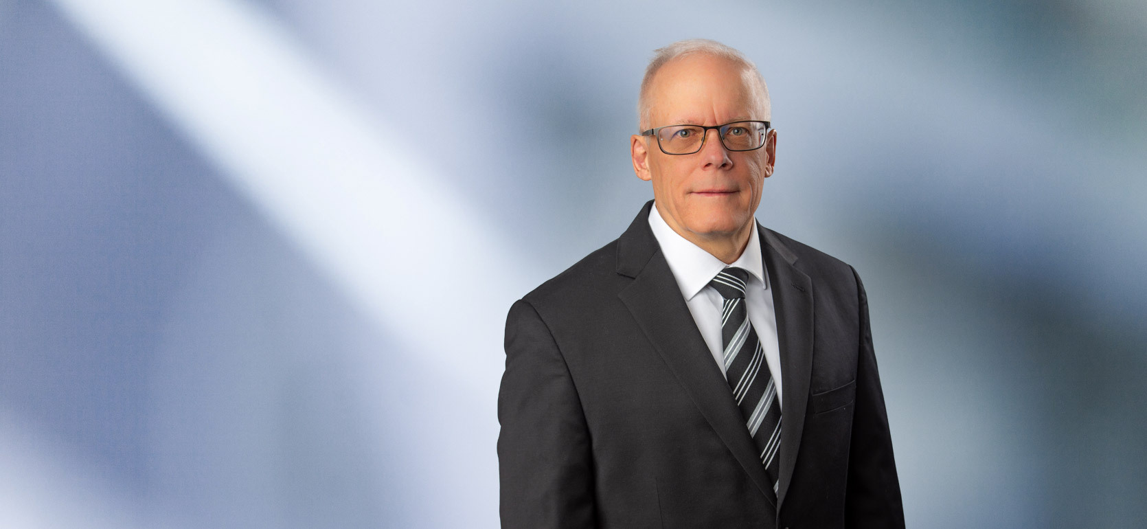 A man, Brent Titus, in a gray suit, light blue shirt, and red patterned tie is looking at the camera, standing against a blurred blue and white background.