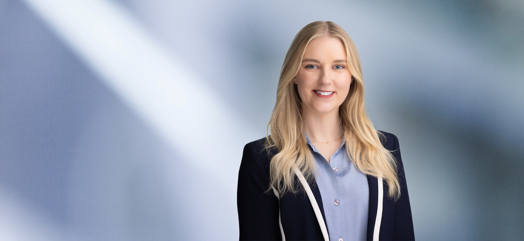 A woman, Alaina Nelson, with long blonde hair wearing a navy blazer over a light blue blouse stands in front of a blurred, blue-toned background.
