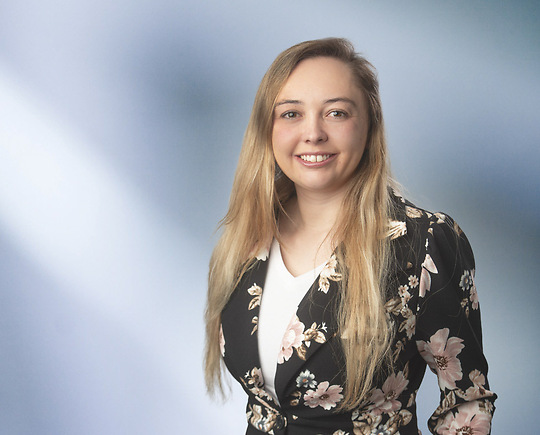 A woman, Jordan Asbury, in a black blazer with a white blouse and a floral pattern is smiling at the camera, standing against a blurred blue and white background.