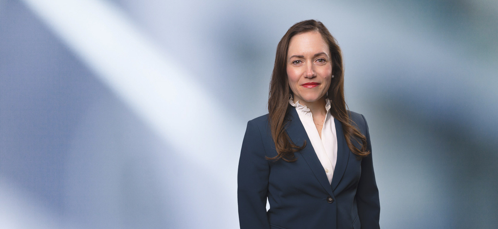 A woman, Gabrielle Lawrence, in a dark suit, light shirt, smiling at the camera, standing against a blurred blue and white background.