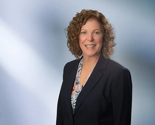 Smiling woman, Cheryl Coutchie, with curly, shoulder-length hair, wearing a navy blazer over a floral blouse, standing in front of a soft blue gradient background.