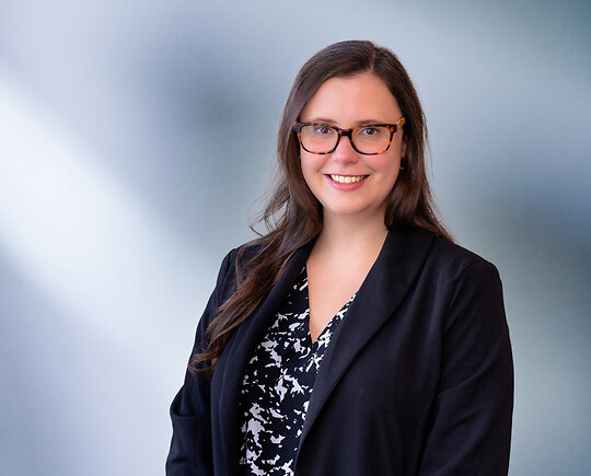 A brown-haired woman, Amy Marinkovski, wearing turtle frame glasses, black sport coat and black and white zigzag pattern shirt, smiles at camera in front of blue gray background