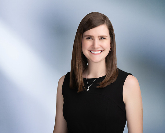 A woman, Amanda Dernovshek, in a sleeveless black dress is smiling at the camera, standing against a blurred blue and white background.