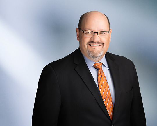 A man, Kevin Roragen, in a black suit, light blue shirt, and orange patterned tie is smiling at the camera, standing against a blurred blue and white background.