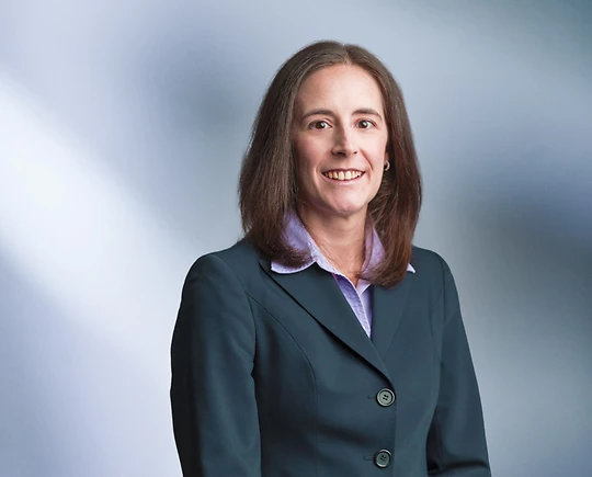 A woman, Erica Huddas, in a dark green blazer and light purple blouse is smiling at the camera, standing against a blurred blue and white background.