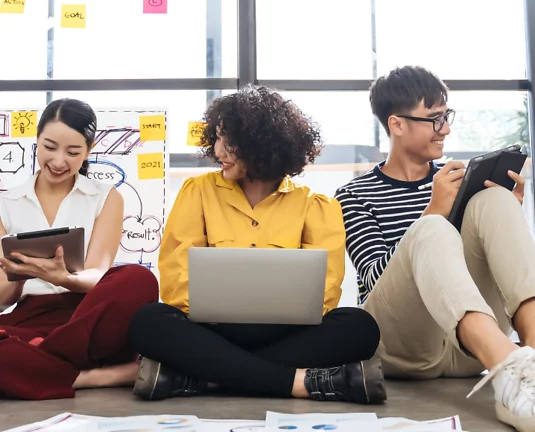 Three young professionals sitting on the floor in a modern office space; one is reading a book, one is using a laptop, and one is holding a tablet. Sticky notes and charts are visible in the background, suggesting a collaborative brainstorming session.