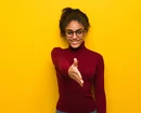 Confident young woman wearing glasses and a red turtleneck offering a handshake while smiling, standing against a bright yellow background.
