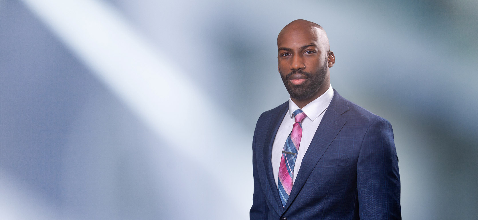 Bald bearded African American man, Xavier Prather, smiles at camera wearing a blue blazer, white undershirt and pink and blue striped necktie in front of blue gray blurred background.