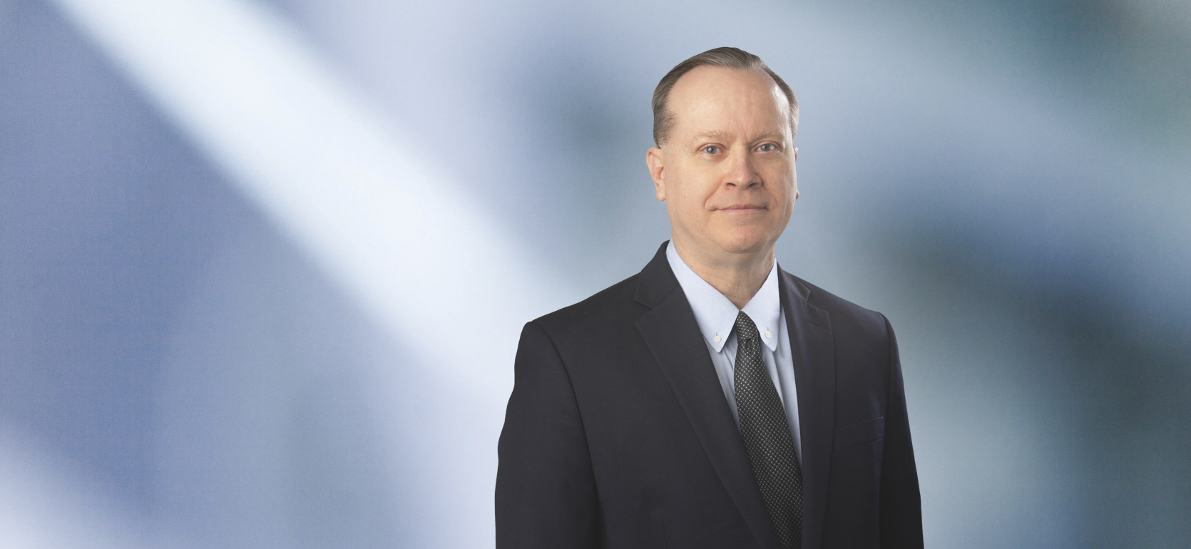 A man, Doug Mielock, in a black suit, light blue shirt, and dark patterned tie is looking at the camera, standing against a blurred blue and white background.