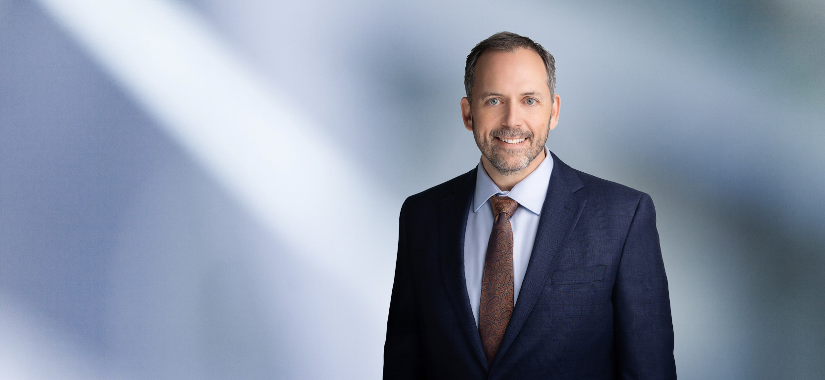 Man, Mikhail Murshak, with short dark hair wearing a navy blue blazer, smiling slightly against a blurred white and blue background.