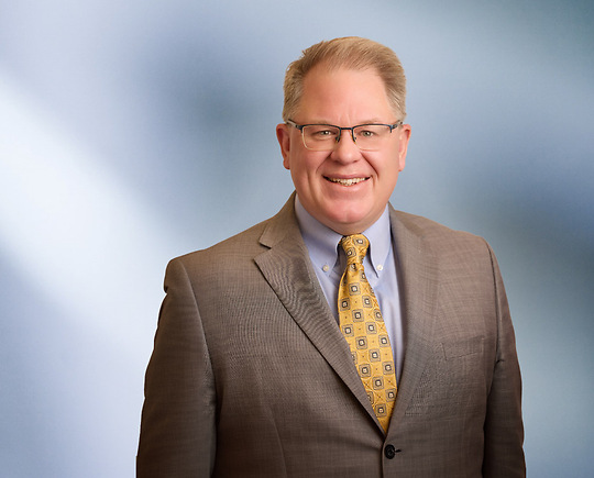 Headshot of a man, Ryan Lamb, with light brown hair, wearing a grey suit and white shirt, smiling against a blurred blue and white background"