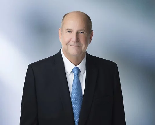 Professional headshot of a middle-aged man, Brian Cermak, in a black suit, white shirt, and light blue tie, standing against a soft blue gradient background.