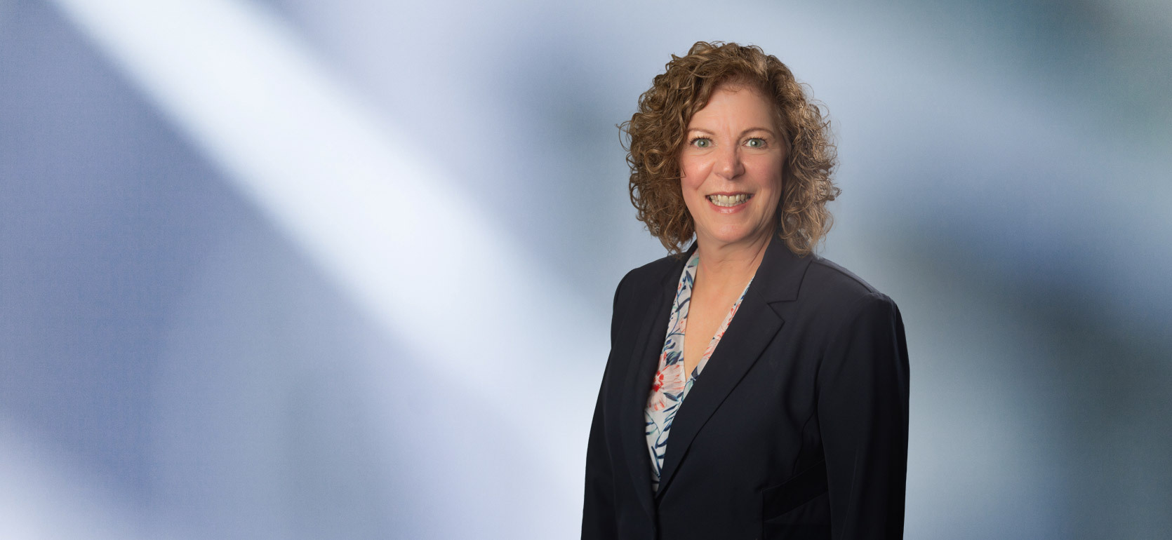 Smiling woman, Cheryl Coutchie, with curly, shoulder-length hair, wearing a navy blazer over a floral blouse, standing in front of a soft blue gradient background.
