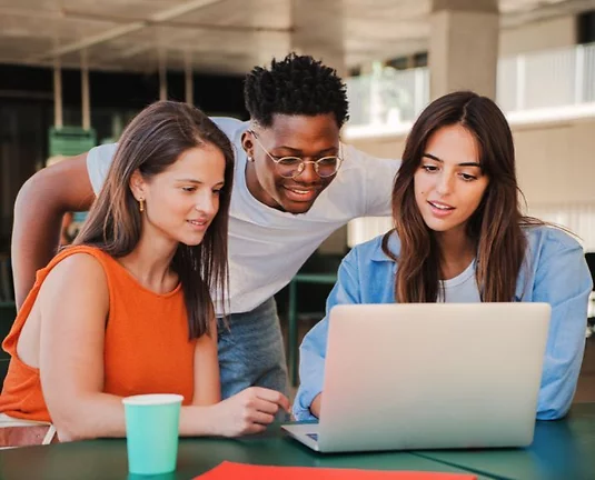 Three college students gathered around a laptop, collaborating and smiling while working on a project. They are seated at a table in a modern indoor-outdoor setting with notebooks and a paper cup nearby.
