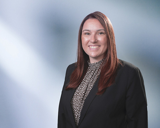 Woman, Stephanie Frizzell, wearing a dark blazer, standing against a blurred blue and white background.