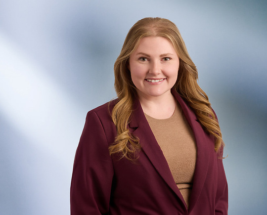 “A woman, Mallory Reader, with shoulder-length, curly blonde hair, wearing a black blazer and white top, is smiling at the camera, standing against a blurred blue and white background.”