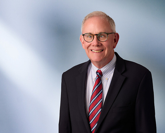 Man, Scott Hogan, wearing a dark blazer, standing against a blurred blue and white background.
