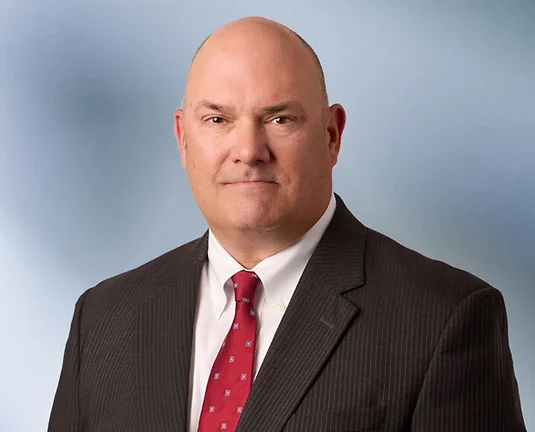 Bald man, Andy Vredenburg looks at camera wearing black pinstripe jacket, white button up shirt and red necktie in front of blurred blue gray background
