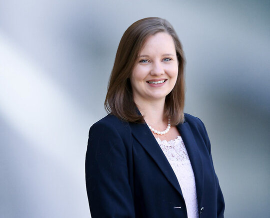Smiling woman, Allison Collins, with shoulder-length light brown hair, wearing a dark blazer, white lace blouse, and pearl necklace, standing in front of a soft blue gradient background