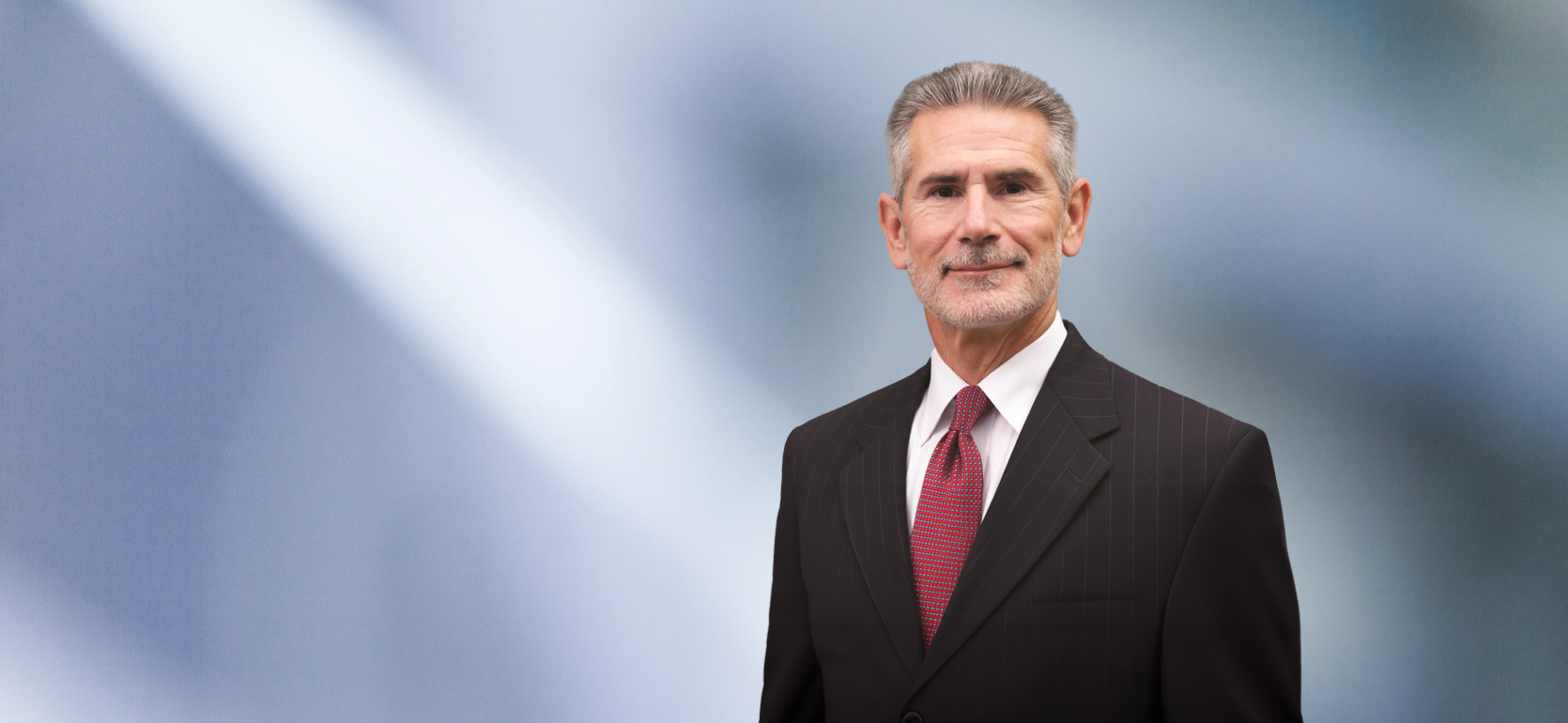 A man, Lawrence Korolewicz, in a dark pinstripe suit, white shirt, and red patterned tie is looking at the camera, standing against a blurred blue and white background.