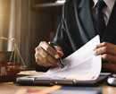 Close-up of a person in a business suit reviewing and signing a document at a desk, with legal scales visible in the background.
