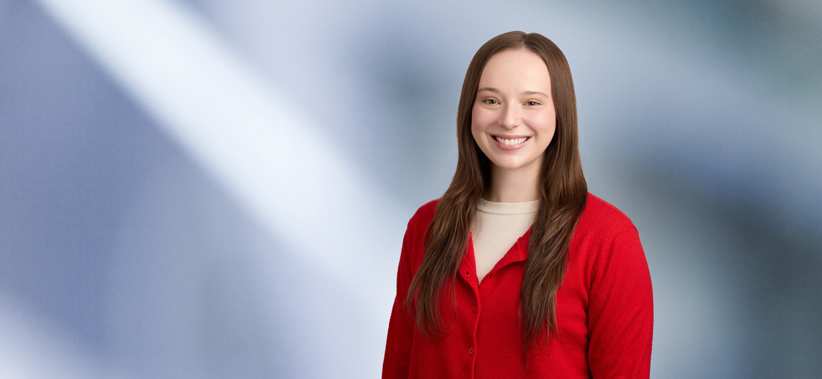 A young woman, Emma Benoit, in a red cardigan and cream top is smiling at the camera, standing against a blurred blue and white background.