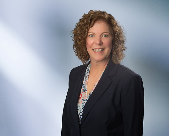 Smiling woman, Cheryl Coutchie, with curly, shoulder-length hair, wearing a navy blazer over a floral blouse, standing in front of a soft blue gradient background.