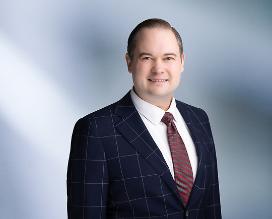 A man, Alexander Rusek, in a dark suit, light shirt, and patterned tie is smiling at the camera, standing against a blurred blue and white background.