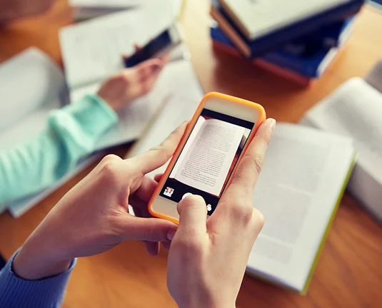 Pair of hands holds cell phone with screen facing towards them as they take screenshots of open book to make cheat sheets