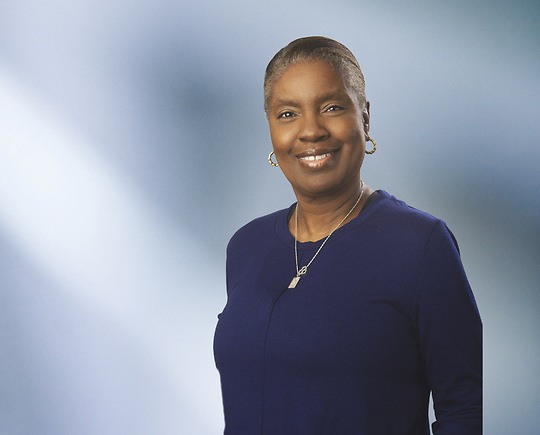 A woman, Gillian Maynard, in a dark blazer and white blouse is smiling at the camera, standing against a blurred blue and white background.