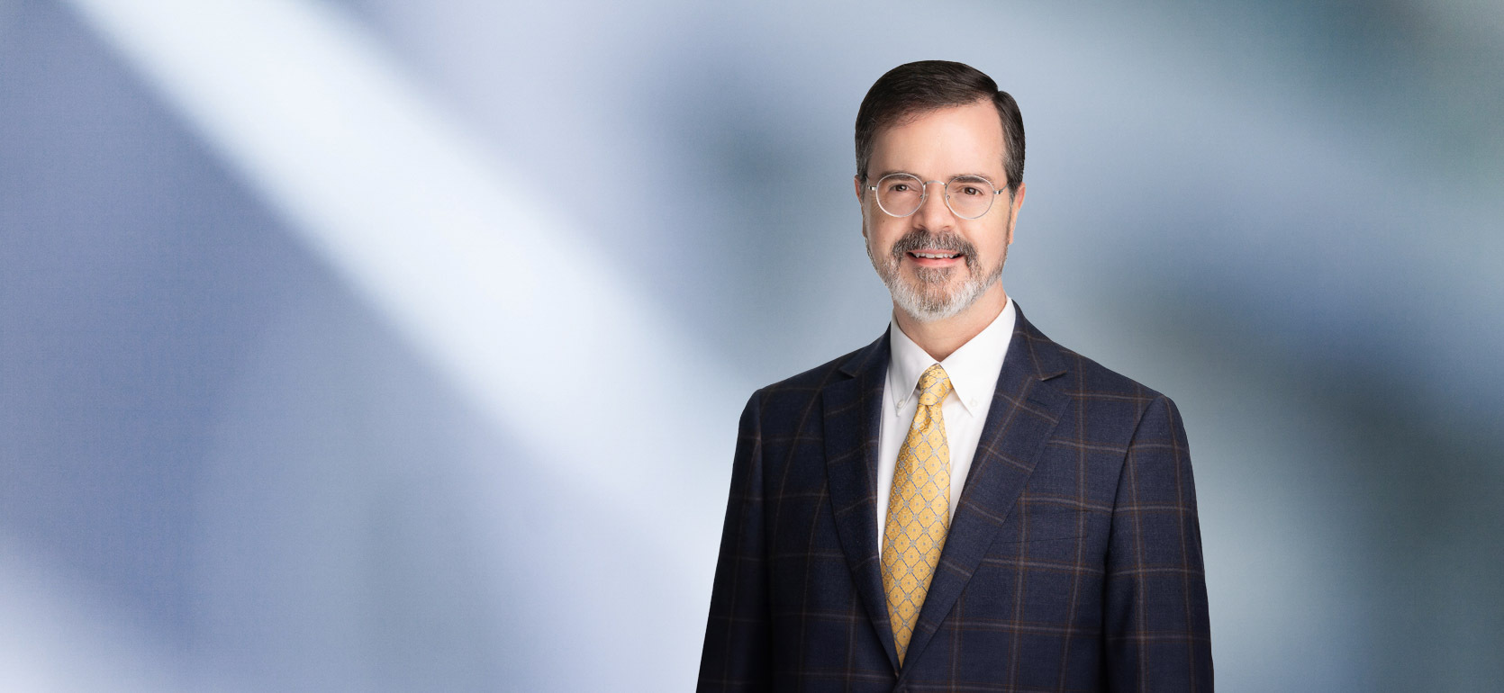 A man, Jeffrey Theuer, in a dark checkered suit, white shirt, and yellow patterned tie is smiling at the camera, standing against a blurred blue and white background.