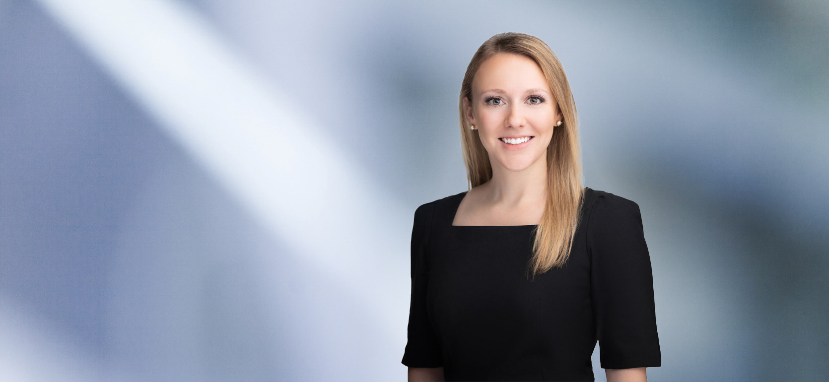 “A woman, McKenna Rievrs, with long, straight dark blonde hair, wearing a black top, is smiling at the camera, standing against a blurred blue and white background.”