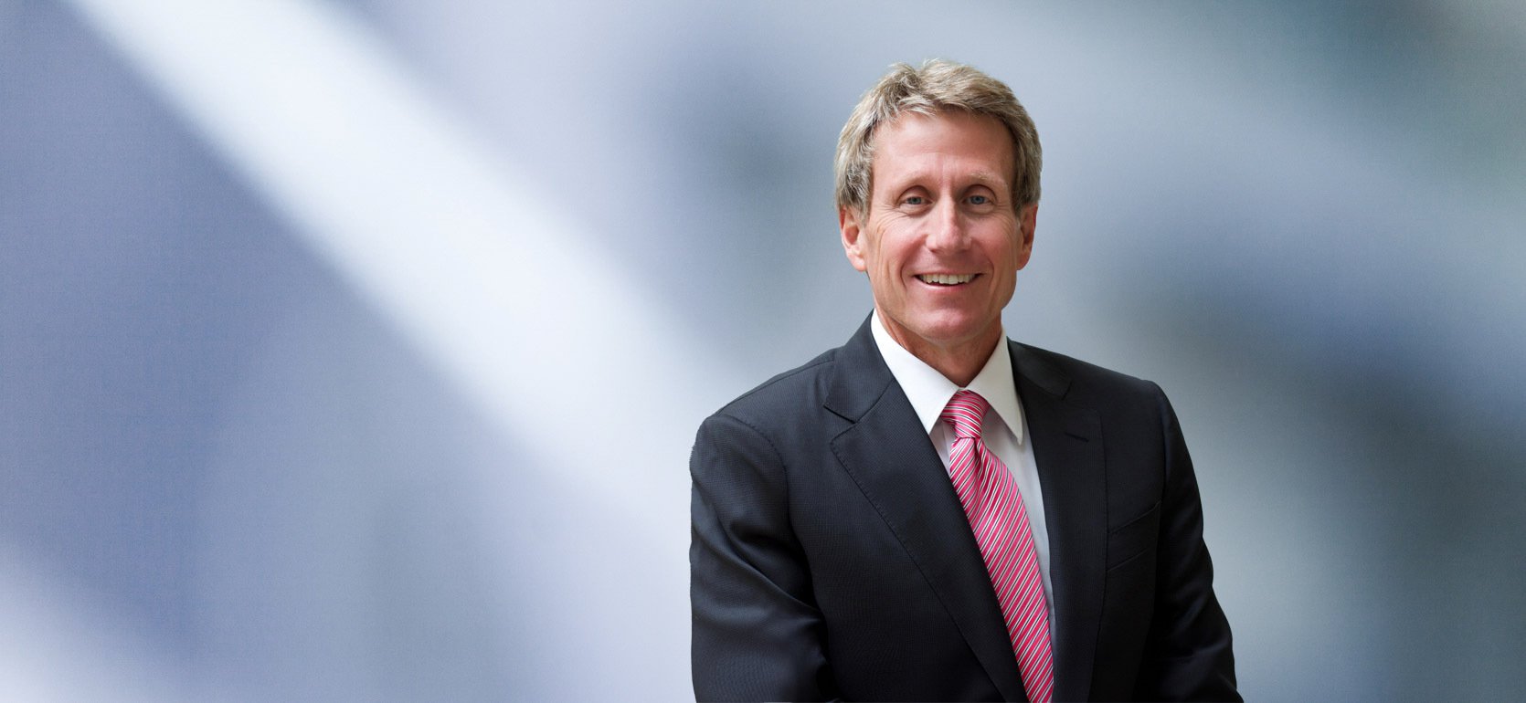 A man, Michael Sanders, with short, dark brown hair, wearing a black blazer, is smiling at the camera, standing against a blurred blue and white background."