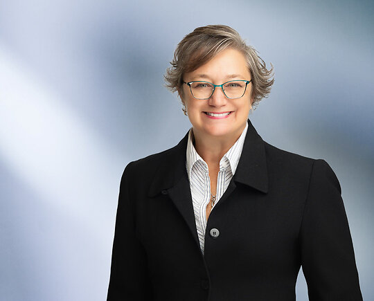 A woman, Heidi Hopper, in a black jacket with white pinstripe shirt and turquoise glasses is smiling at the camera, standing against a blurred blue and white background.