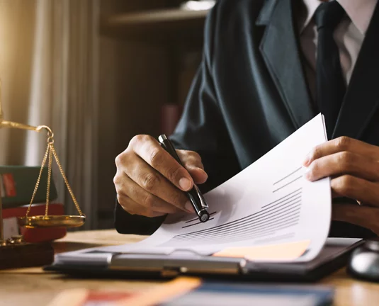 Close-up of a person in a business suit reviewing and signing a document at a desk, with legal scales visible in the background.