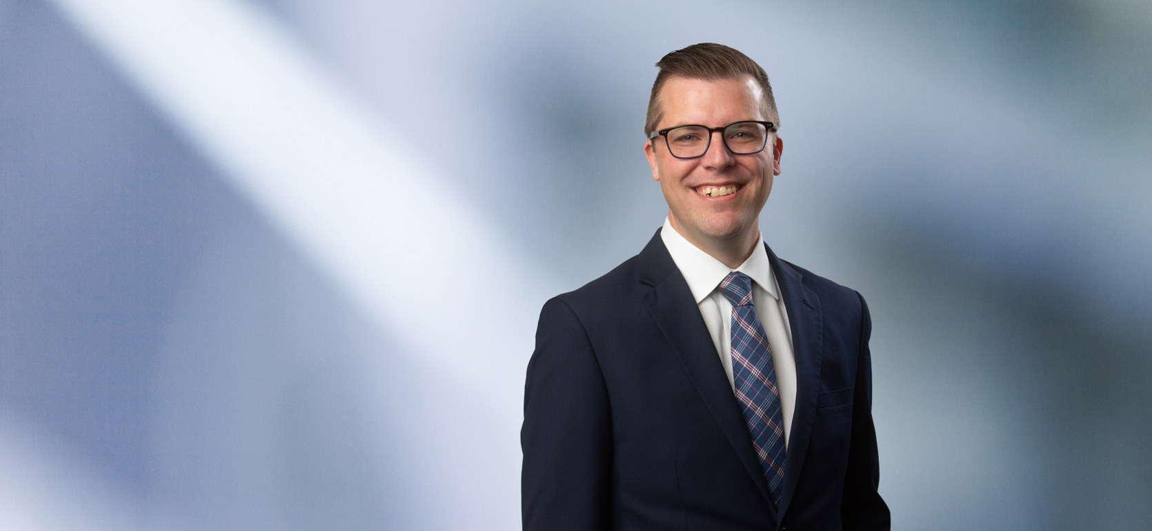 A man, Taylor Gast, in a dark suit smiling at the camera, standing against a blurred blue and white background.