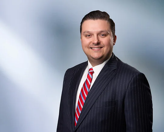 A man, Joe Giacalone, smiles at camera wearing a black pinstripe suit, white shirt and red striped tie in front of blue gray blurred background