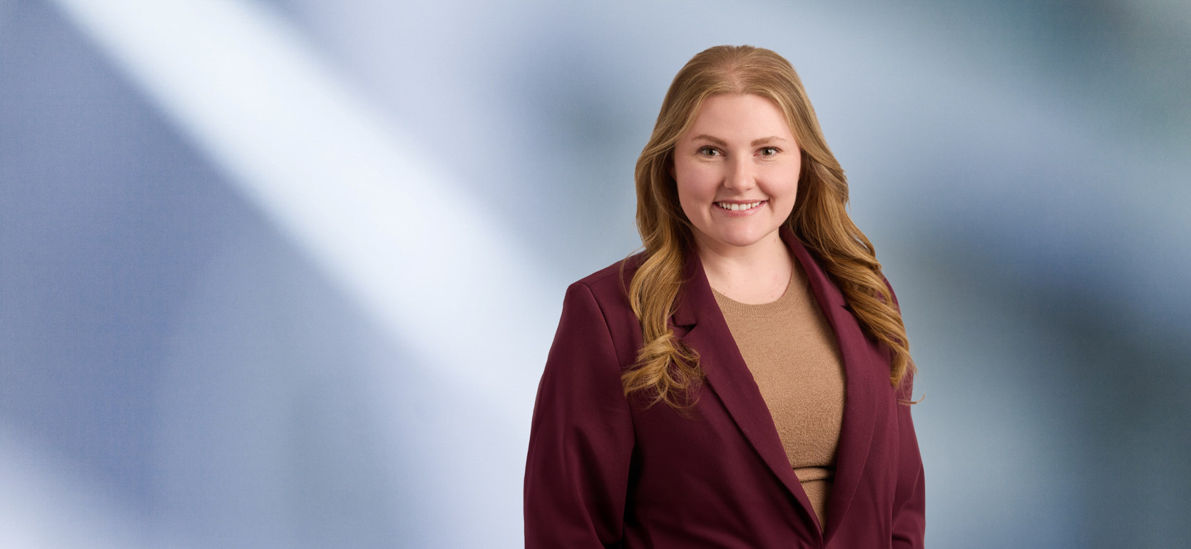“A woman, Mallory Reader, with shoulder-length, curly blonde hair, wearing a black blazer and white top, is smiling at the camera, standing against a blurred blue and white background.”