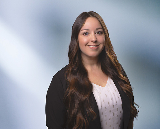 A woman, Shamarr Gearhart, in a dark suit and light blouse is smiling at the camera, standing against a blurred blue and white background.