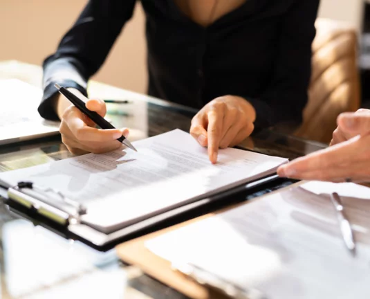 Close-up of two professionals reviewing and discussing a document on a clipboard at a glass desk.