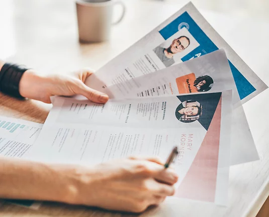Hand holding many paper resumes and writing notes with pen in other hand at desk