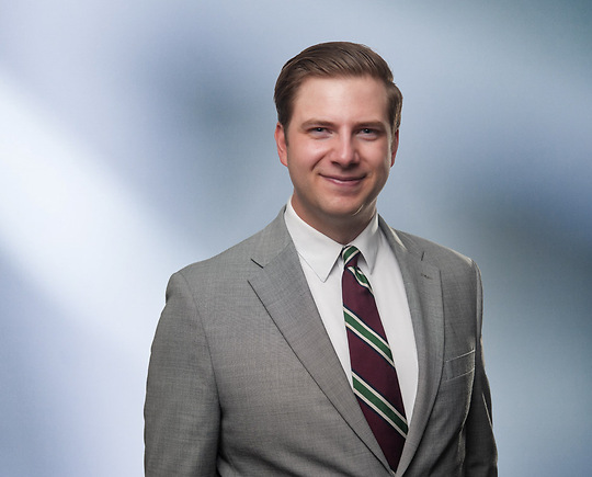 “A man, Matthew Murray, with short, light brown hair, wearing a grey blazer and white shirt, is smiling at the camera, standing against a blurred blue and white background.”