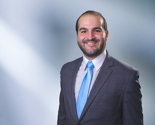 Man, Patrick Quinn, with short brown hair wearing a dark gray blazer, smiling against a blurred white and blue background.
