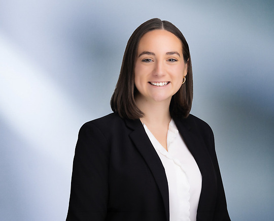 woman, Samantha Shepard, with dark brown hair, wearing a black blazer, smiling against a blurred blue and white background"