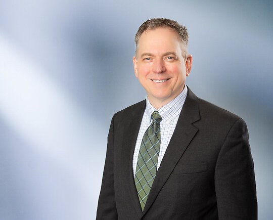 A middle-aged man, Thomas Huyck, smiles at camera, wearking a black sport coat with stripped patterned collared shirt and green necktie in front of blue gray blurred background