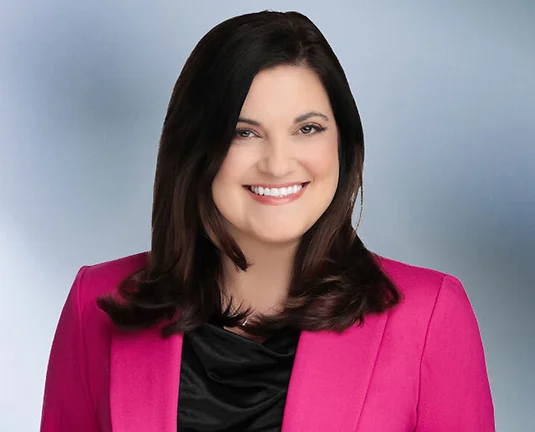 Woman attorney, Stefania Gismondi, smiles at camera in front of blue gray blurred background wearing pink sport coat and black blouse