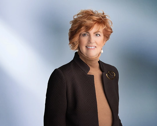 A woman, Kelly Lucas, in a brown textured blazer over a tan turtleneck, accessorized with pearl earrings and a brooch, is smiling at the camera, standing against a blurred blue and white background.