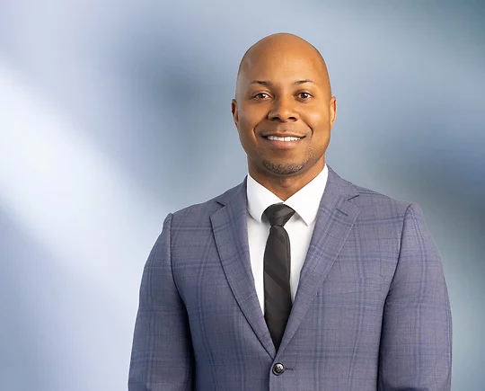 A bald African American man, Ray Littleton, wearing a blue tweed pattern sport coat with a white undershirt and black striped tie is smiling at the camera, standing against a blurred blue and white background.